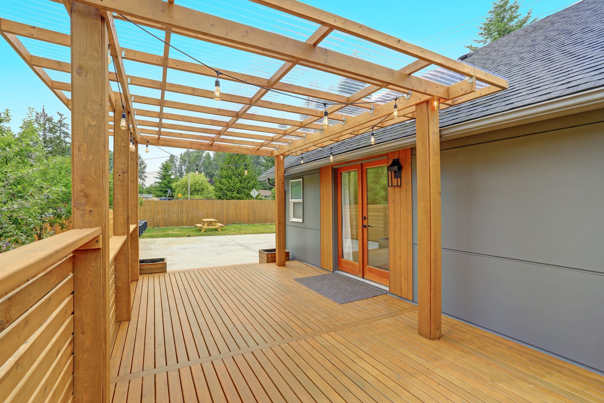 Wooden deck with pergola beside a house, featuring sliding glass doors and a railing overlooking a yard.