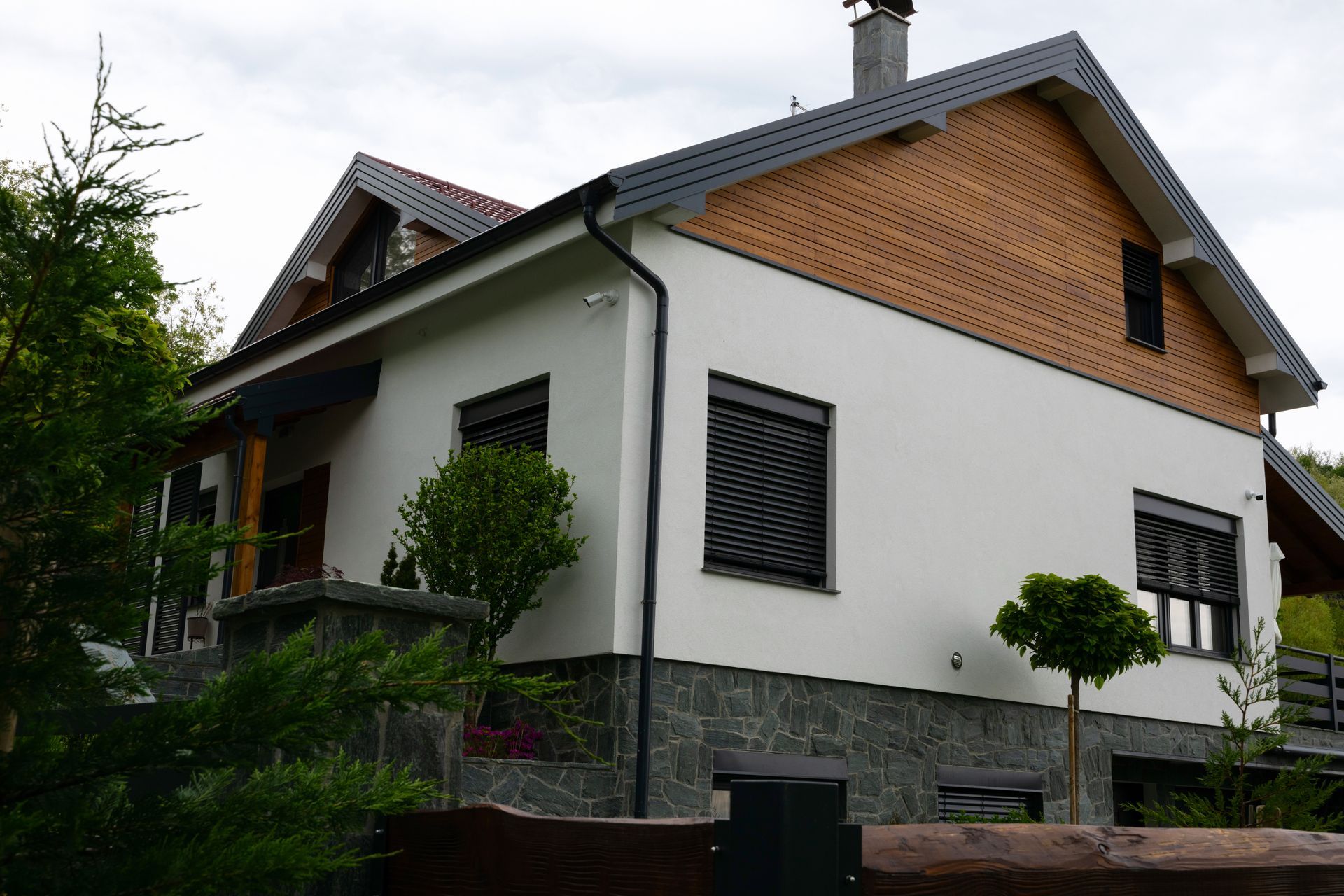 Two-story house with white walls, gray stone base, and orange brick gable roof surrounded by greenery