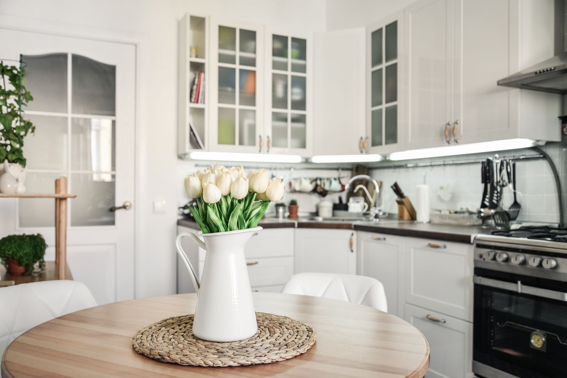 Bright white kitchen with round table, vase of white tulips, and black stove under glass cabinets