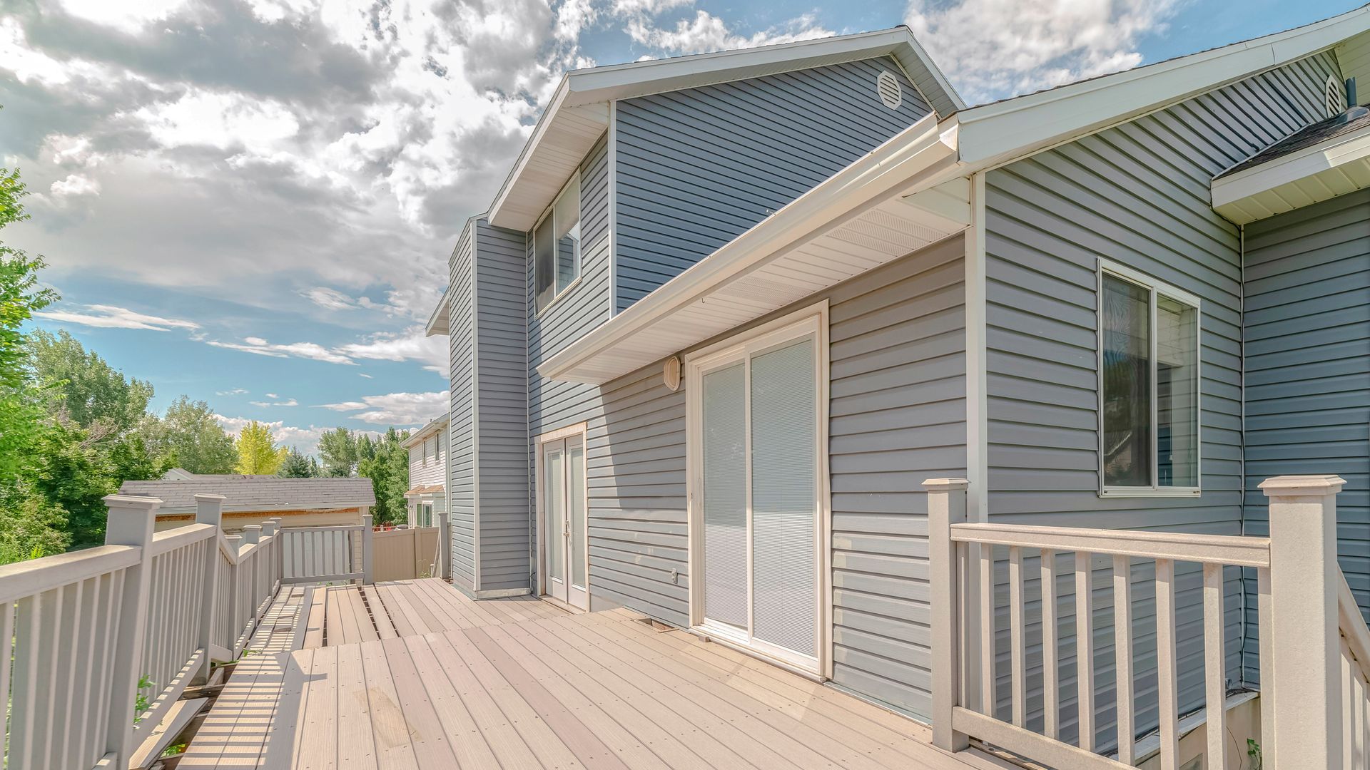 Blue-gray house with a wooden deck, railings, and sliding glass doors under a partly cloudy sky