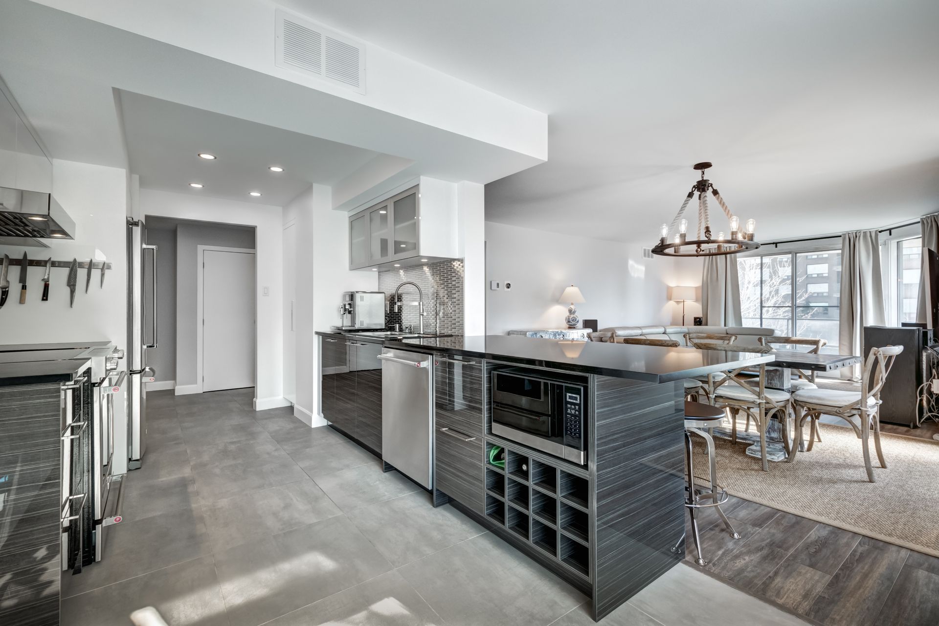 Modern open-concept kitchen and dining area with gray flooring, island wine rack, and pendant chandelier Modern open-concept kitchen and dining area with gray flooring, island wine rack, and pendant chandelier