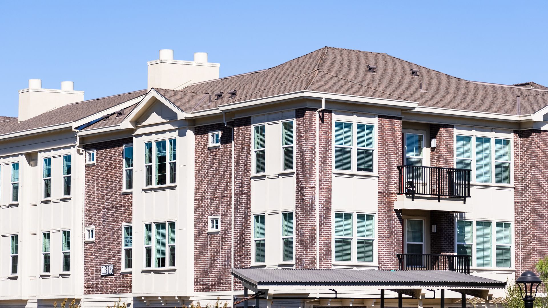 Three-story beige apartment building with brown roof, white-trimmed windows, and balconies under a clear blue sky