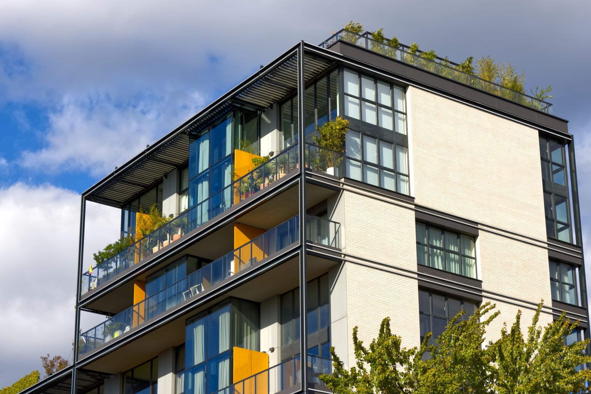 Modern apartment building with glass balconies, yellow accents, and greenery against a blue sky