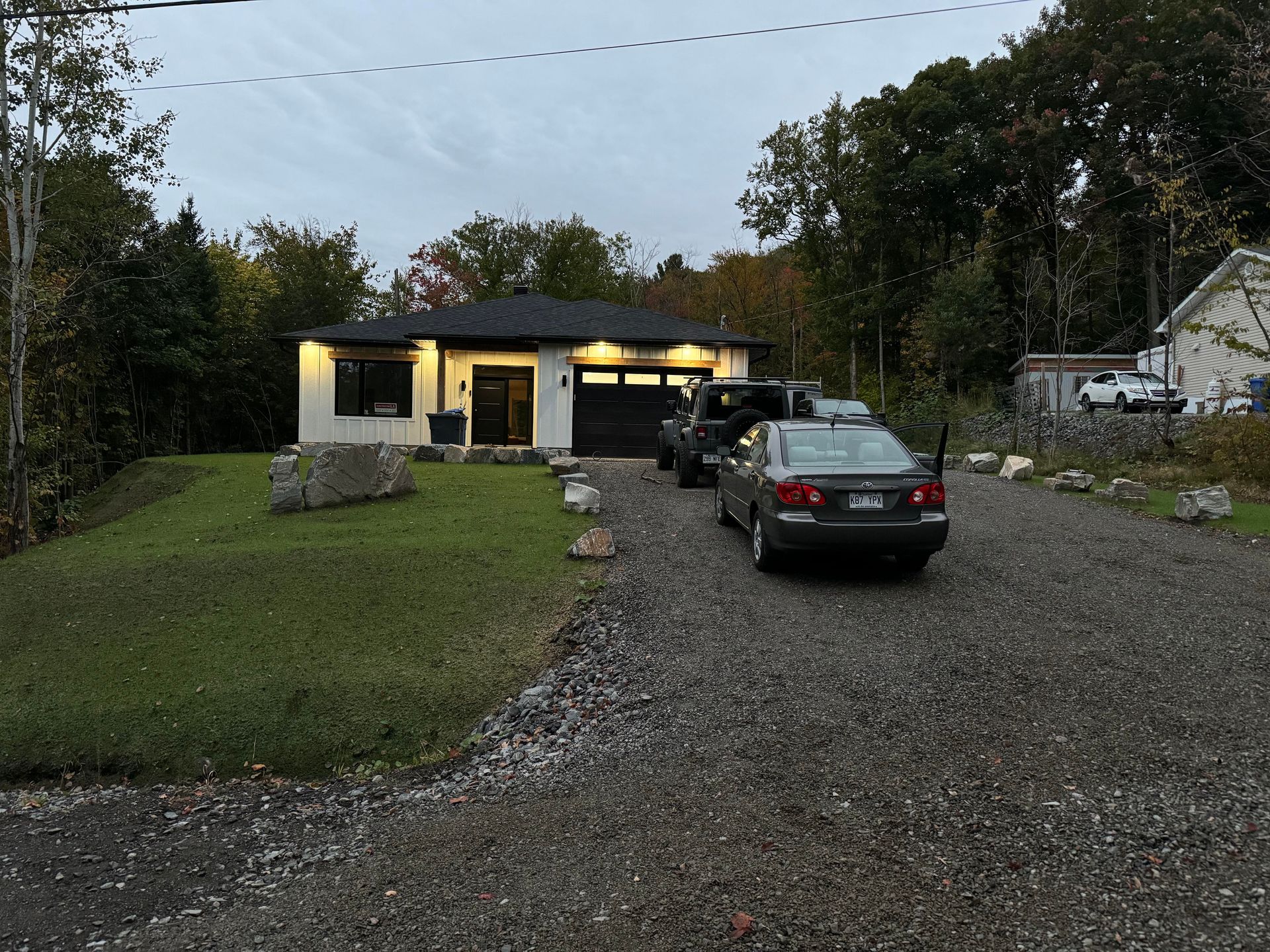 Une petite maison avec une porte de garage et une allée noires, deux voitures garées et une pelouse verte.