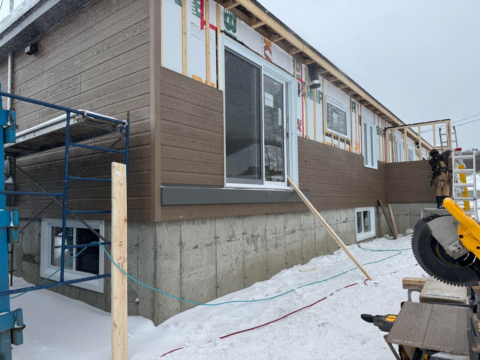 Façade d'une maison en construction, avec un bardage brun, une porte coulissante et de la neige au sol.