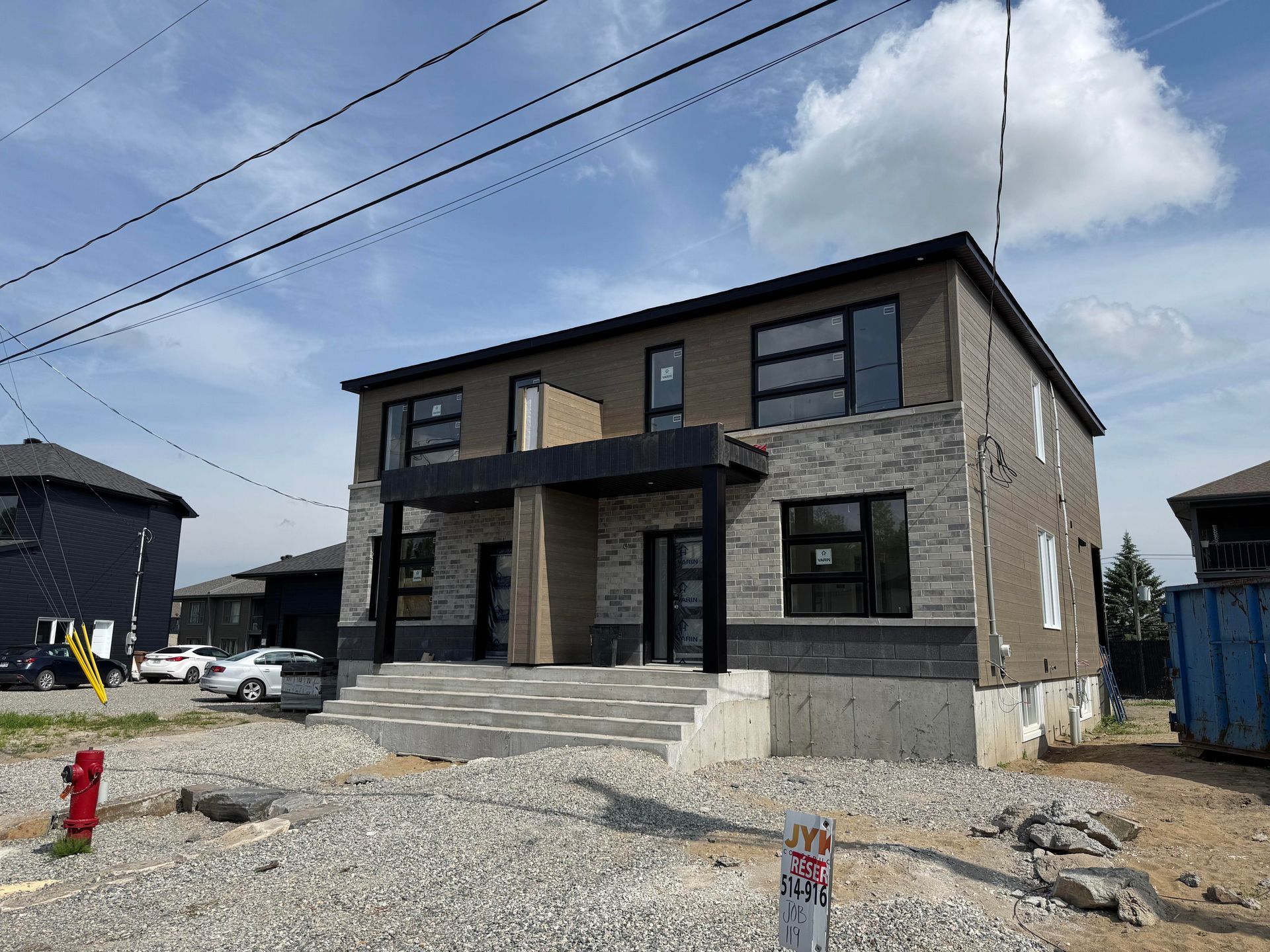 Duplex de deux étages en construction, avec des accents de pierre et de noir, sur un terrain de gravier sous un ciel nuageux.