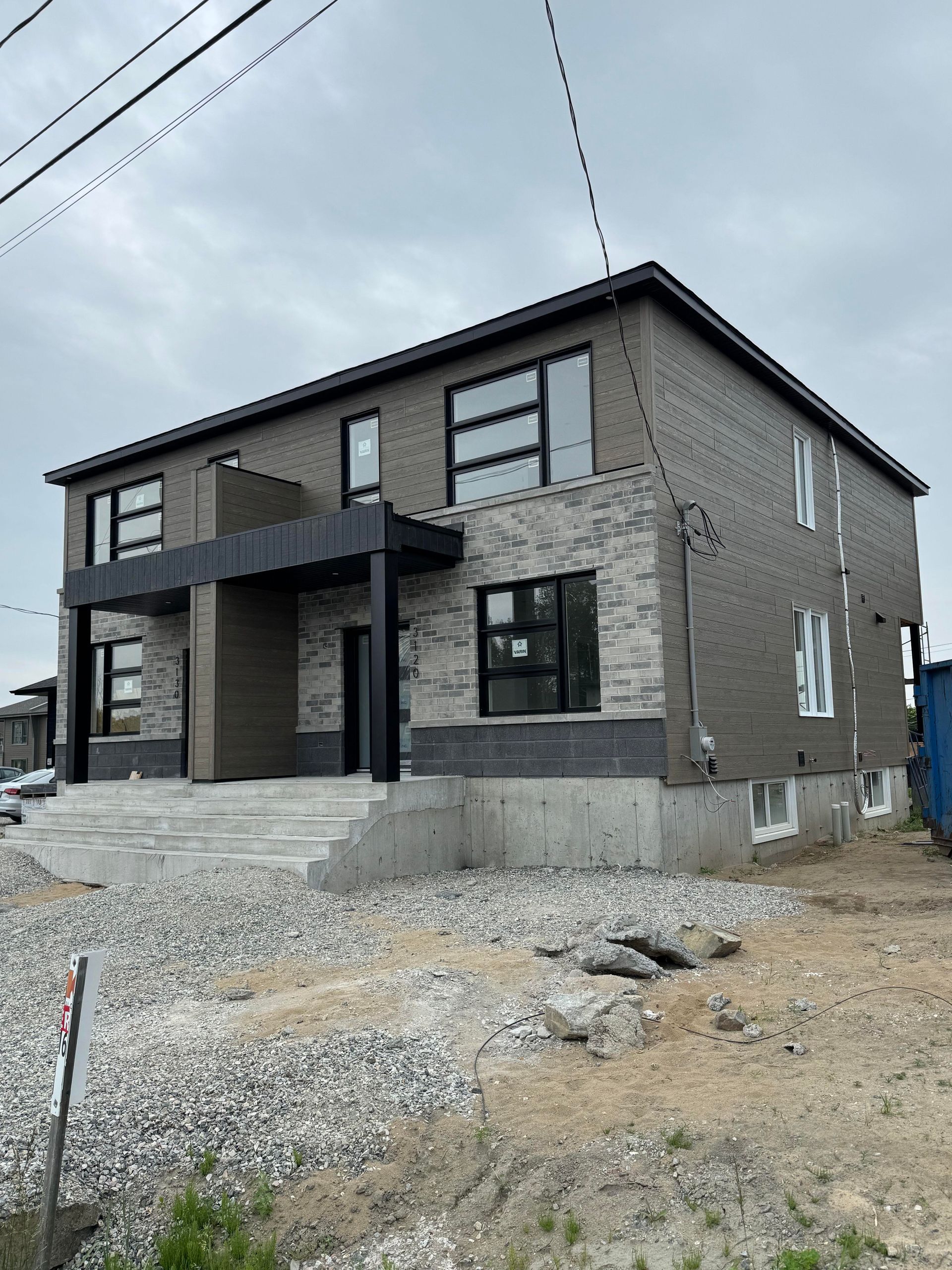 Duplex moderne de deux étages en construction, avec briques grises et fenêtres noires. Gravier et débris de construction devant.