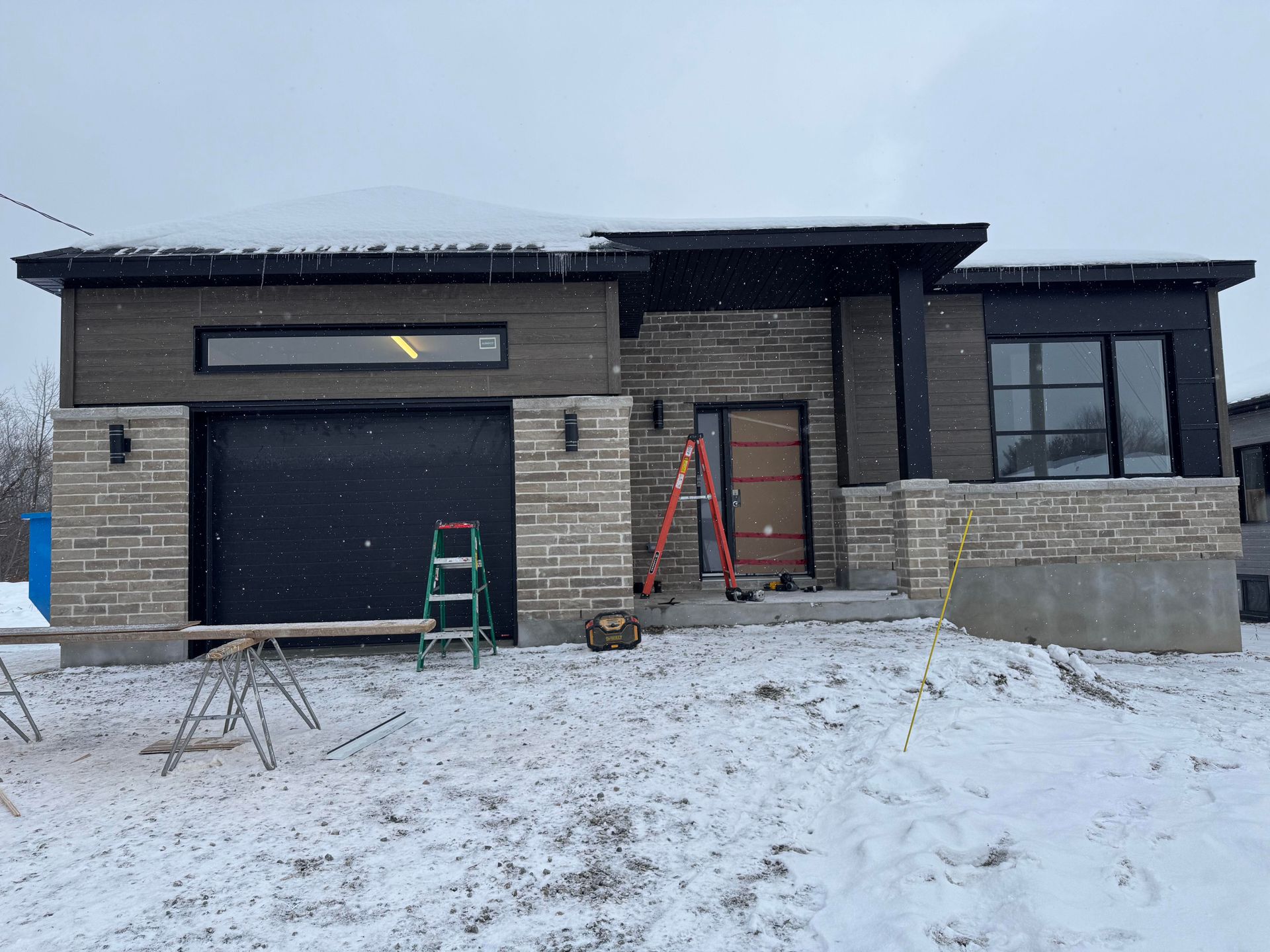 Une maison moderne en construction, avec des murs en pierre et un bardage sombre, de la neige sur le toit et au sol, et des outils de construction.