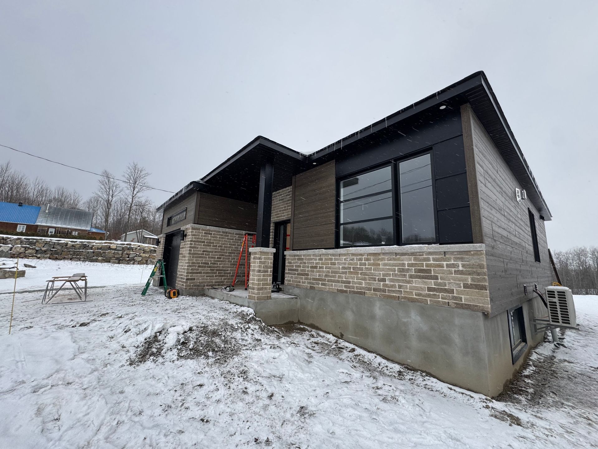 Une maison moderne en construction, avec des façades en pierre et un bardage sombre, se dresse dans un paysage enneigé.