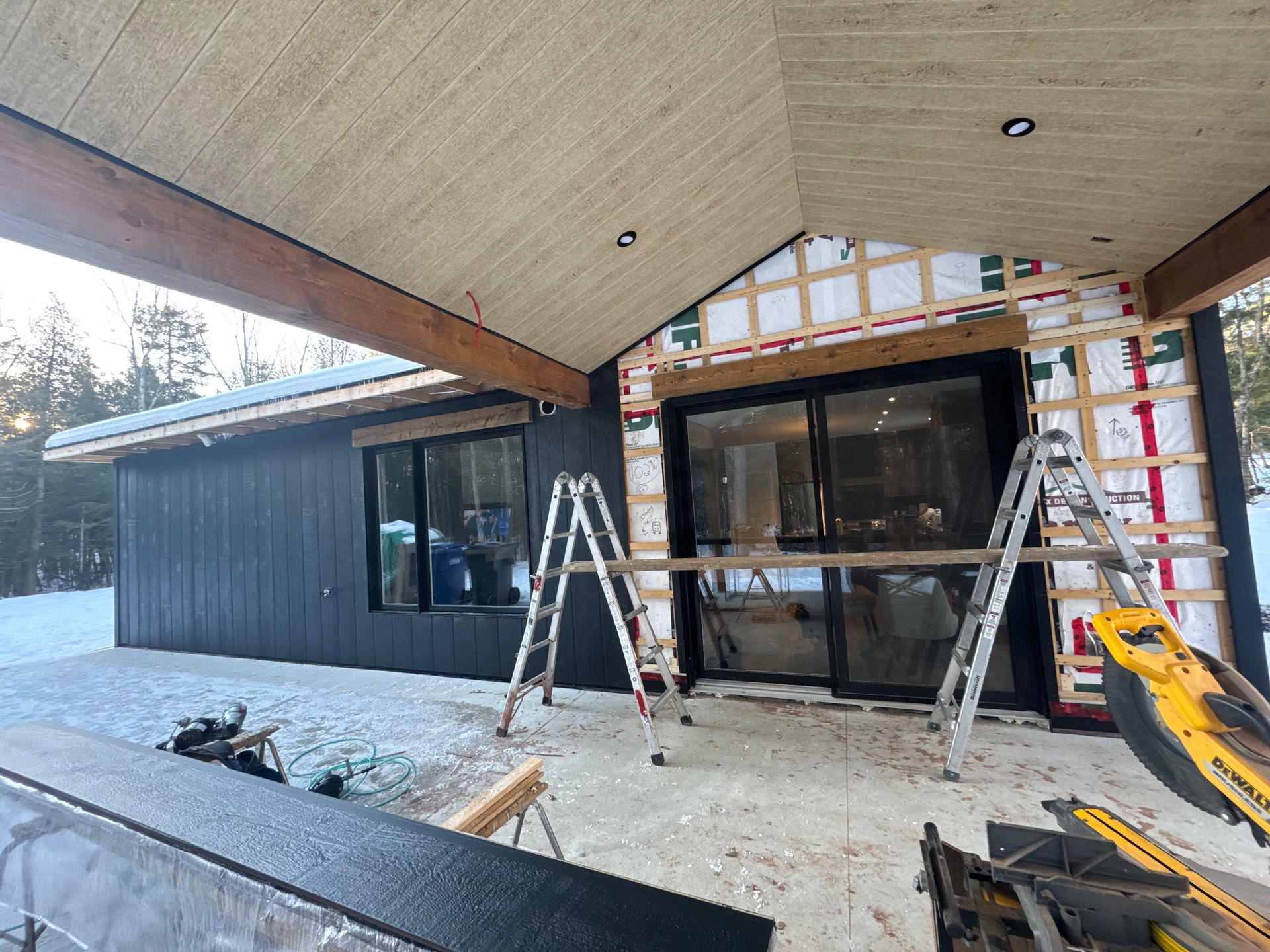 Extérieur d'une maison en construction avec bardage noir, portes coulissantes en verre et isolation visible.