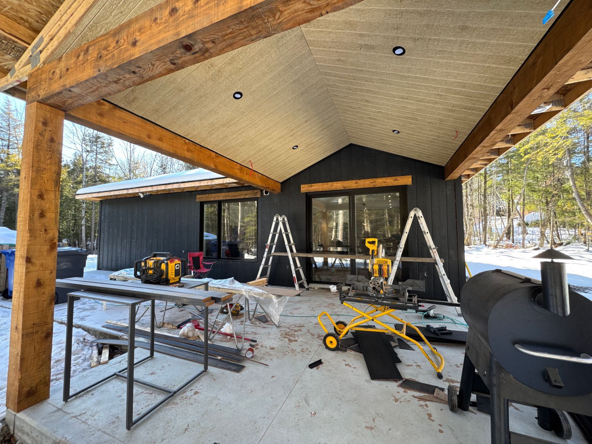 Une cabane gris foncé avec une véranda en construction. Des outils et un fumoir sont présents. Le sol est enneigé.