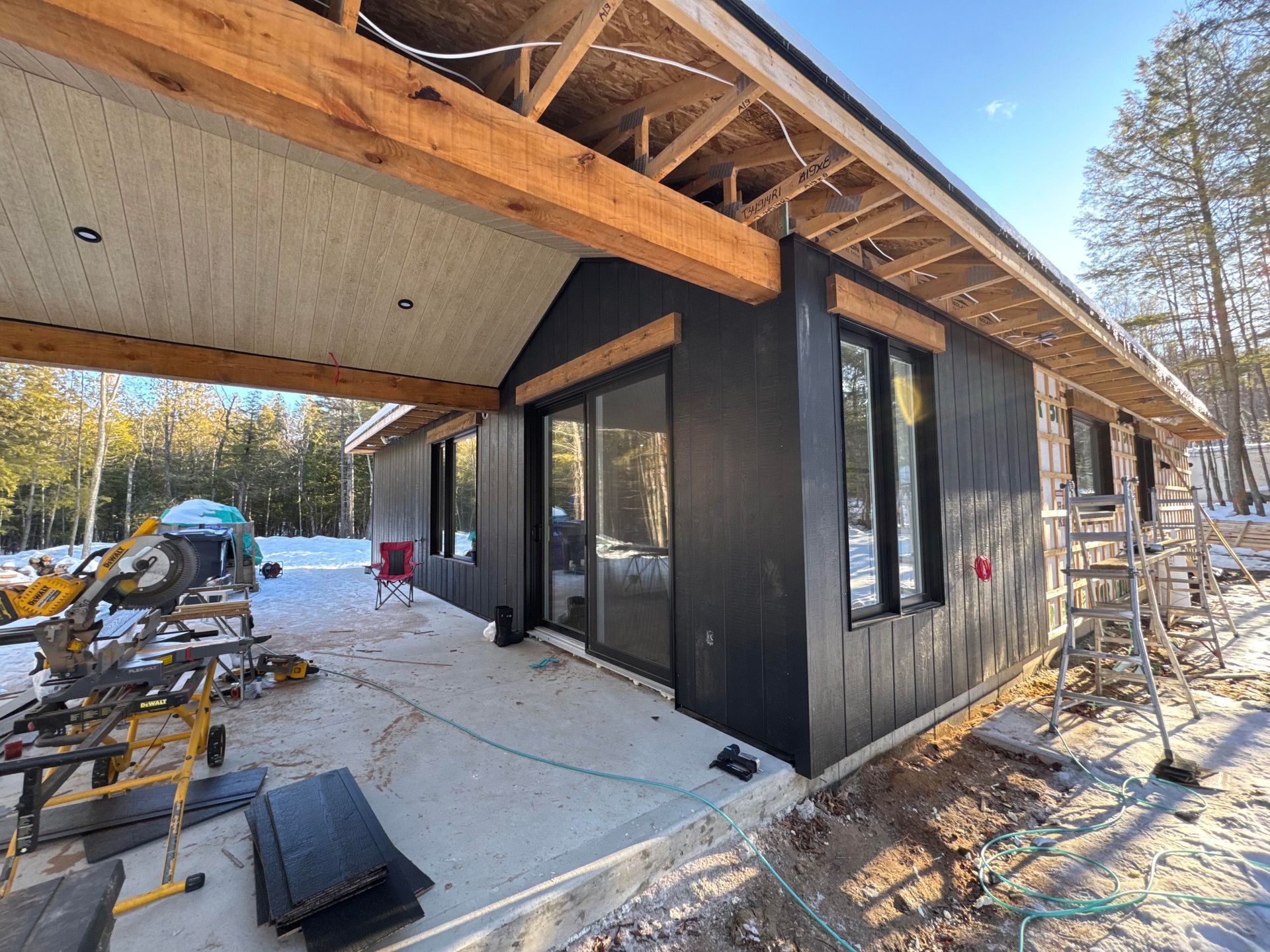 Vue extérieure d'une maison en construction avec un bardage noir, des poutres en bois et une porte coulissante en verre.