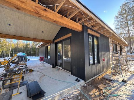 Vue extérieure d'une maison en construction avec un bardage noir, des poutres en bois et une porte coulissante en verre.
