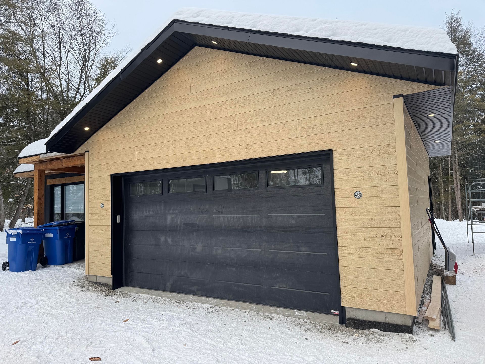 Garage avec porte noire, bardage beige et neige au sol.
