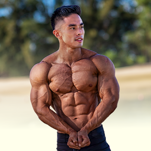 Andee Teo without a shirt is standing on a beach with his hands behind his back.