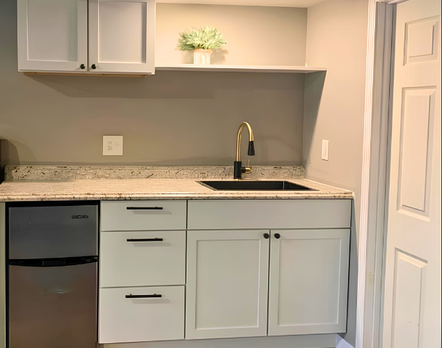Small kitchenette with white cabinets, granite countertop, sink, and black mini fridge under open shelving.