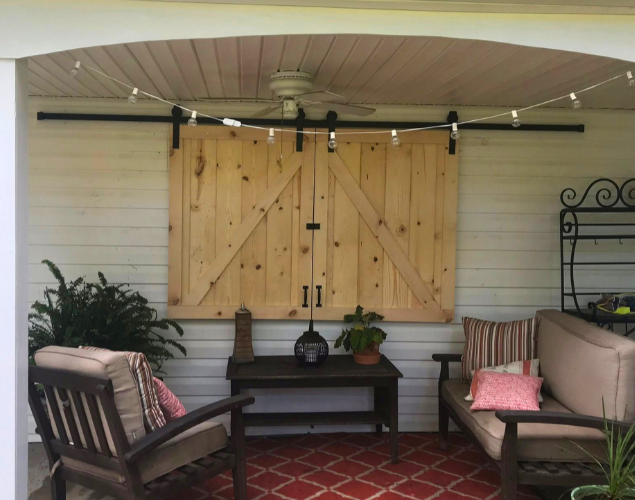 Covered porch with wooden shutters, cushioned chairs, potted plants, and a red patterned rug