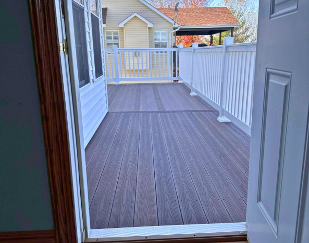 Open doorway to a small deck with brown flooring and white railings, facing neighboring houses.
