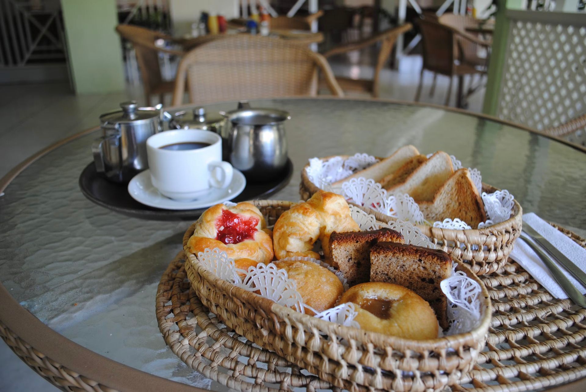 Breakfast tray with pastries, fruit, coffee cups, and a teapot on a glass table