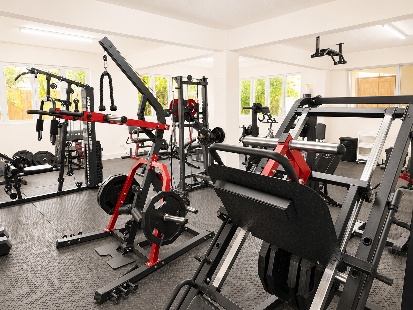 Home gym with red and black exercise machines in a bright room with large windows.