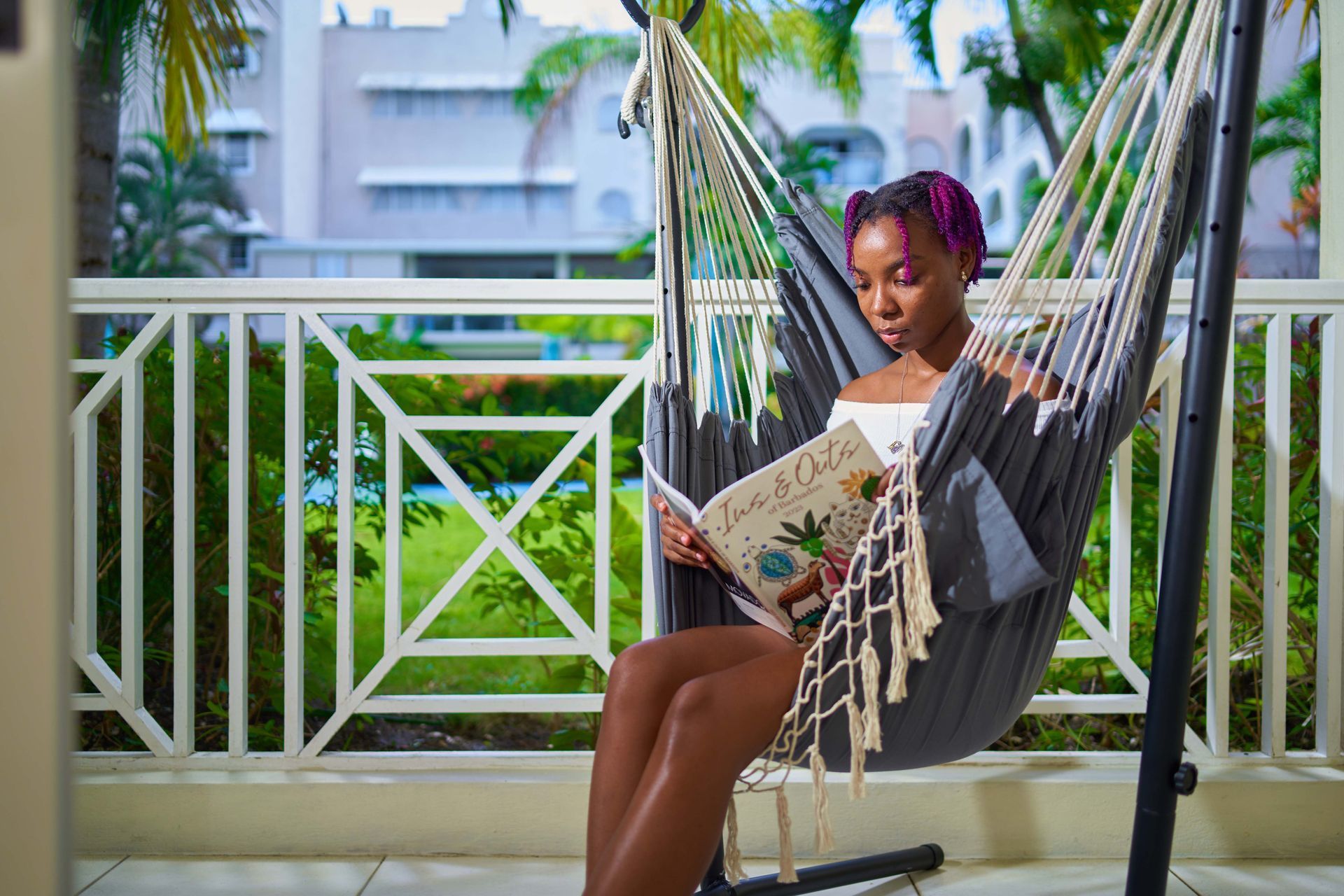 Woman reading in a hammock on a balcony, with tropical plants and a white railing in the background.