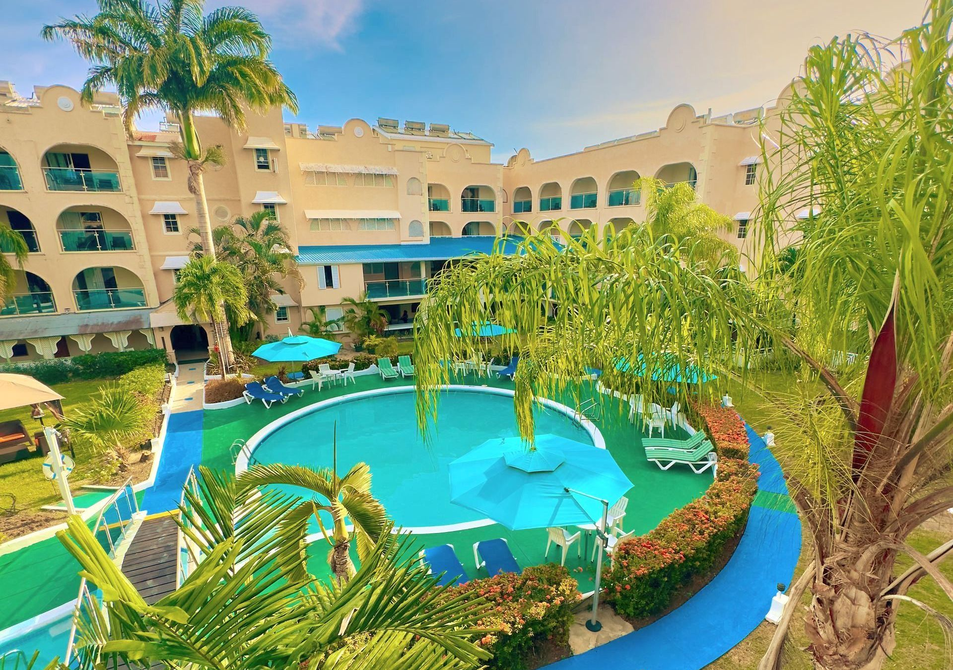 Resort courtyard with turquoise pool, palm trees, and beige balconies under a blue sky