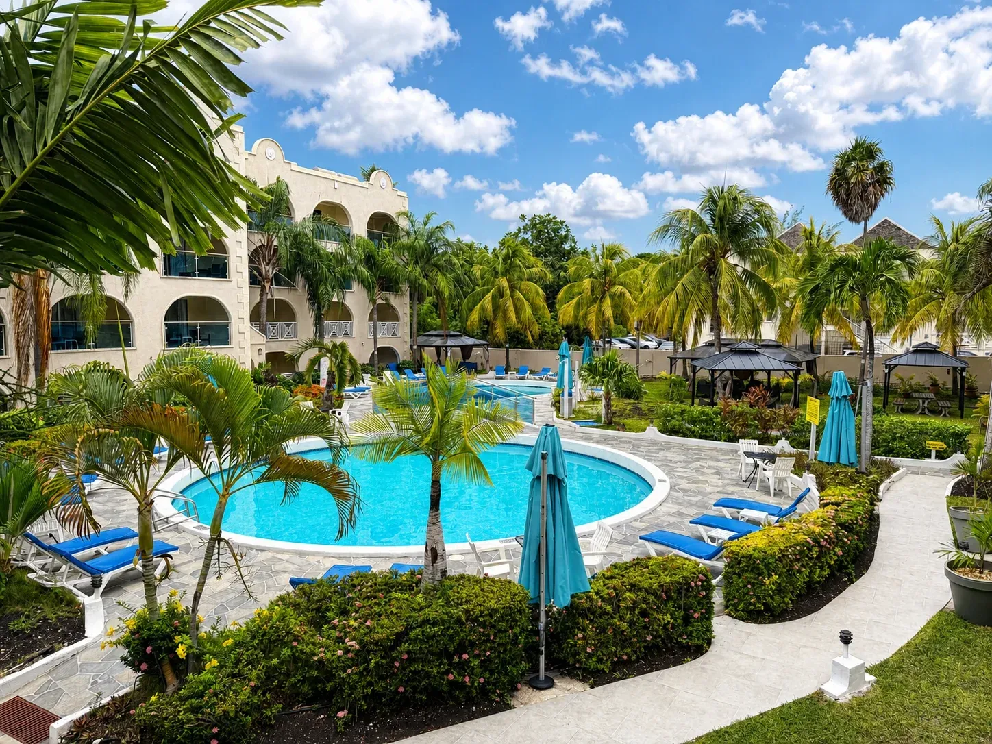 Resort pool with turquoise water, blue umbrellas, palm trees, and white lounge chairs under a sunny sky