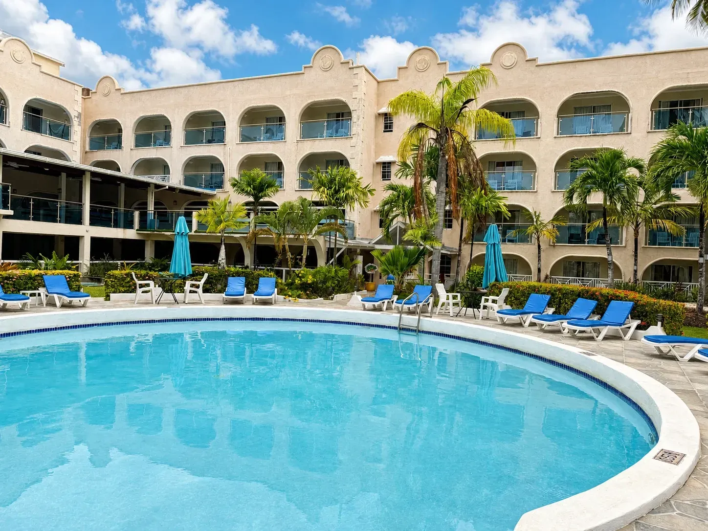 Resort pool with blue lounge chairs, palm trees, and a multi-story hotel under a sunny sky