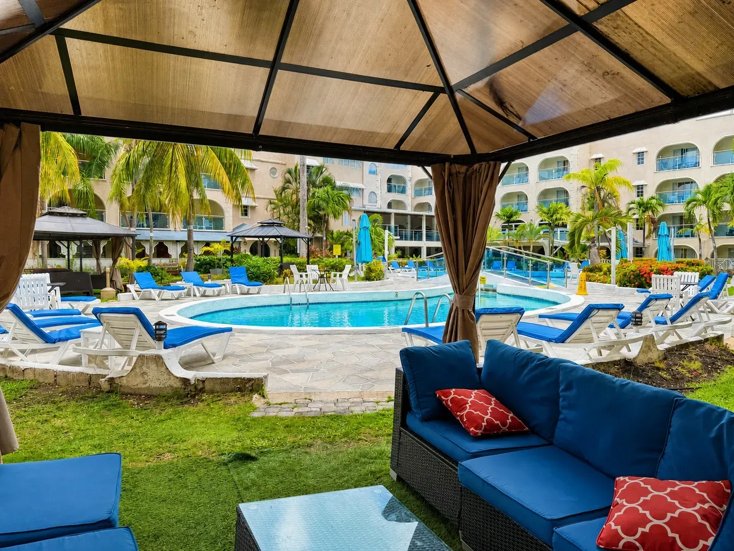 Resort pool view from shaded cabana with blue lounge chairs and a blue sofa with red pillows