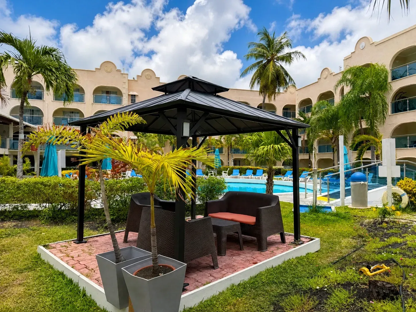 Resort courtyard with a thatched gazebo, pool, palm trees, and cloudy blue sky
