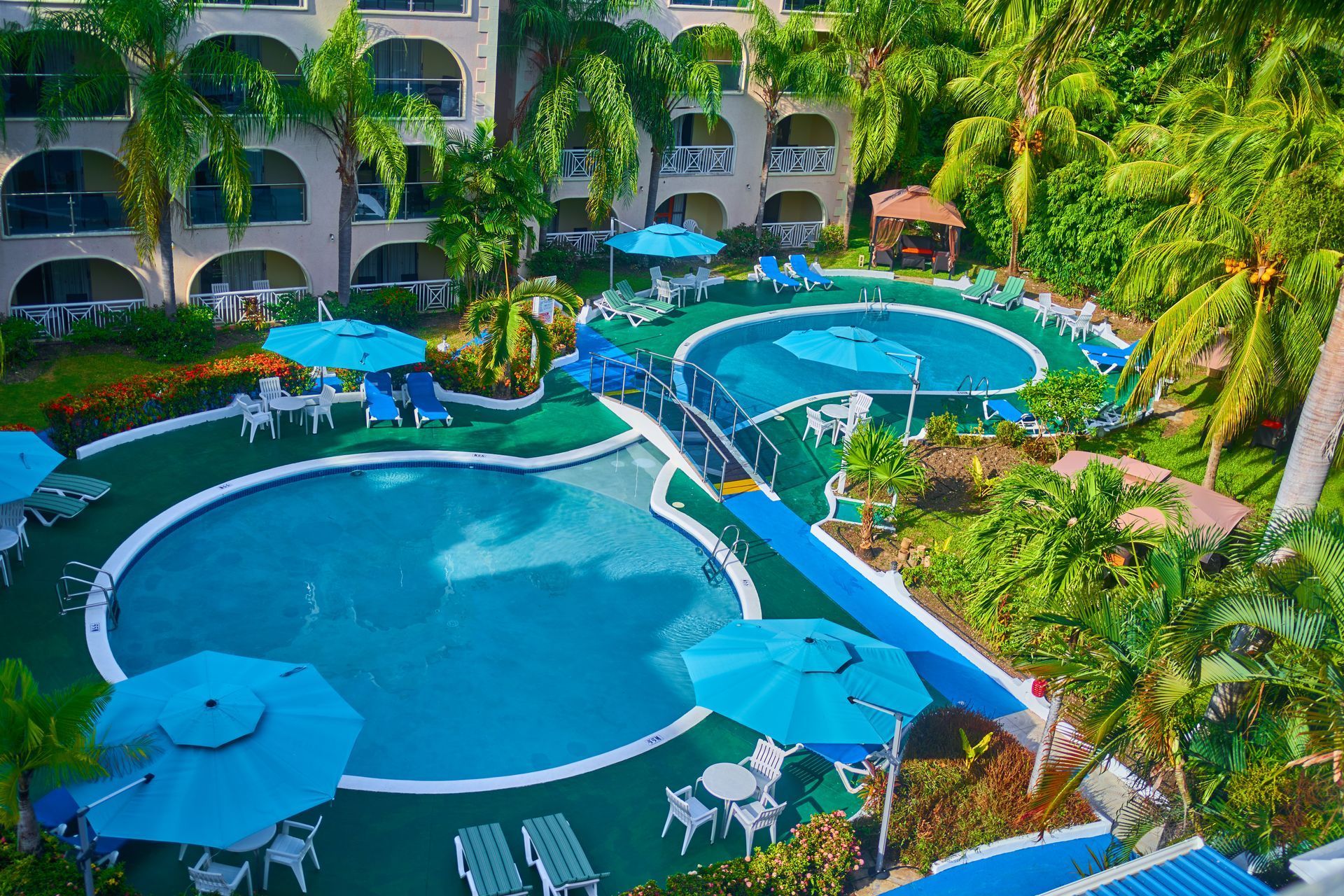 Resort courtyard with multiple blue swimming pools, white umbrellas, palm trees, and surrounding balconies