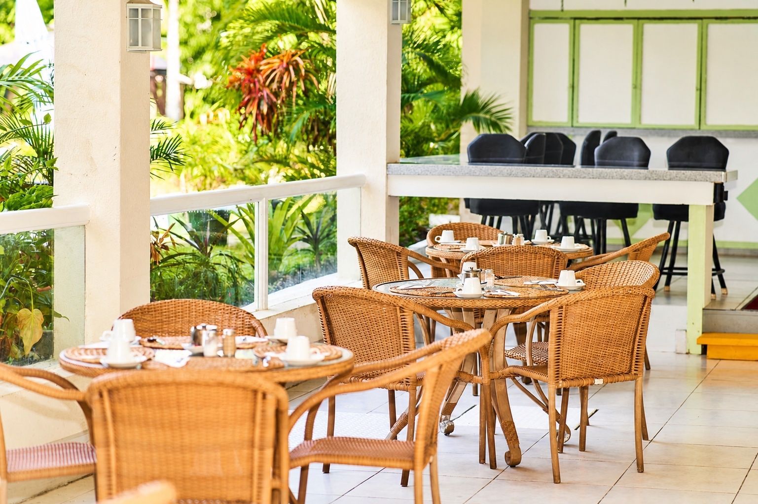 Outdoor café patio with wicker chairs, set tables, and tropical greenery beside a black grill table