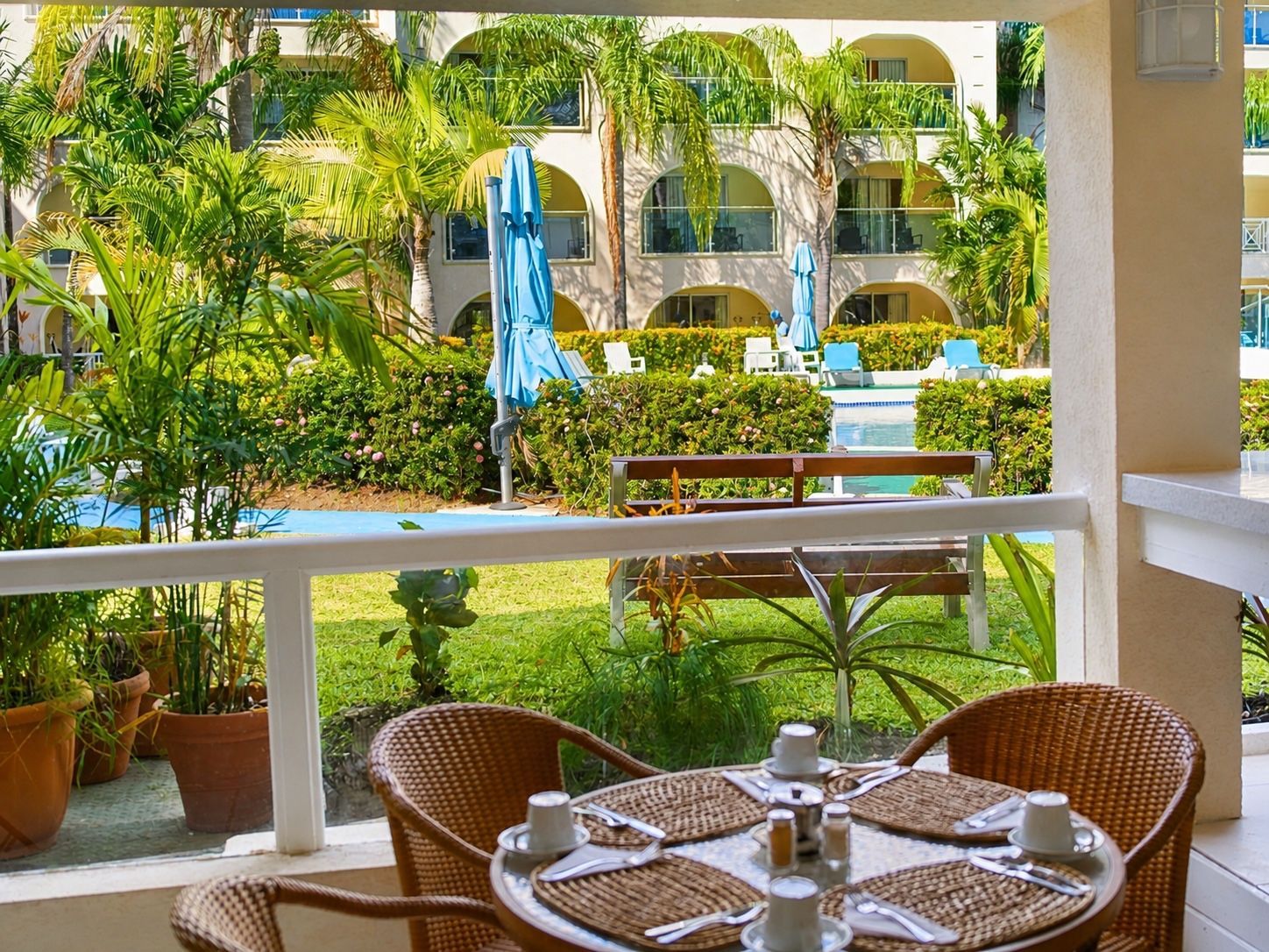 Patio dining area with wicker chairs, set table, lush garden, and blue fountain in a sunny courtyard