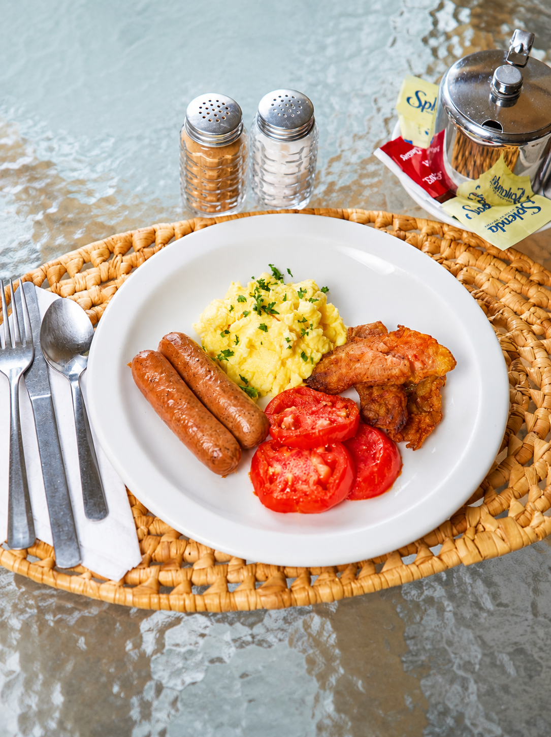 Breakfast plate with sausages, scrambled eggs, tomatoes, and fried chicken on a woven placemat by water
