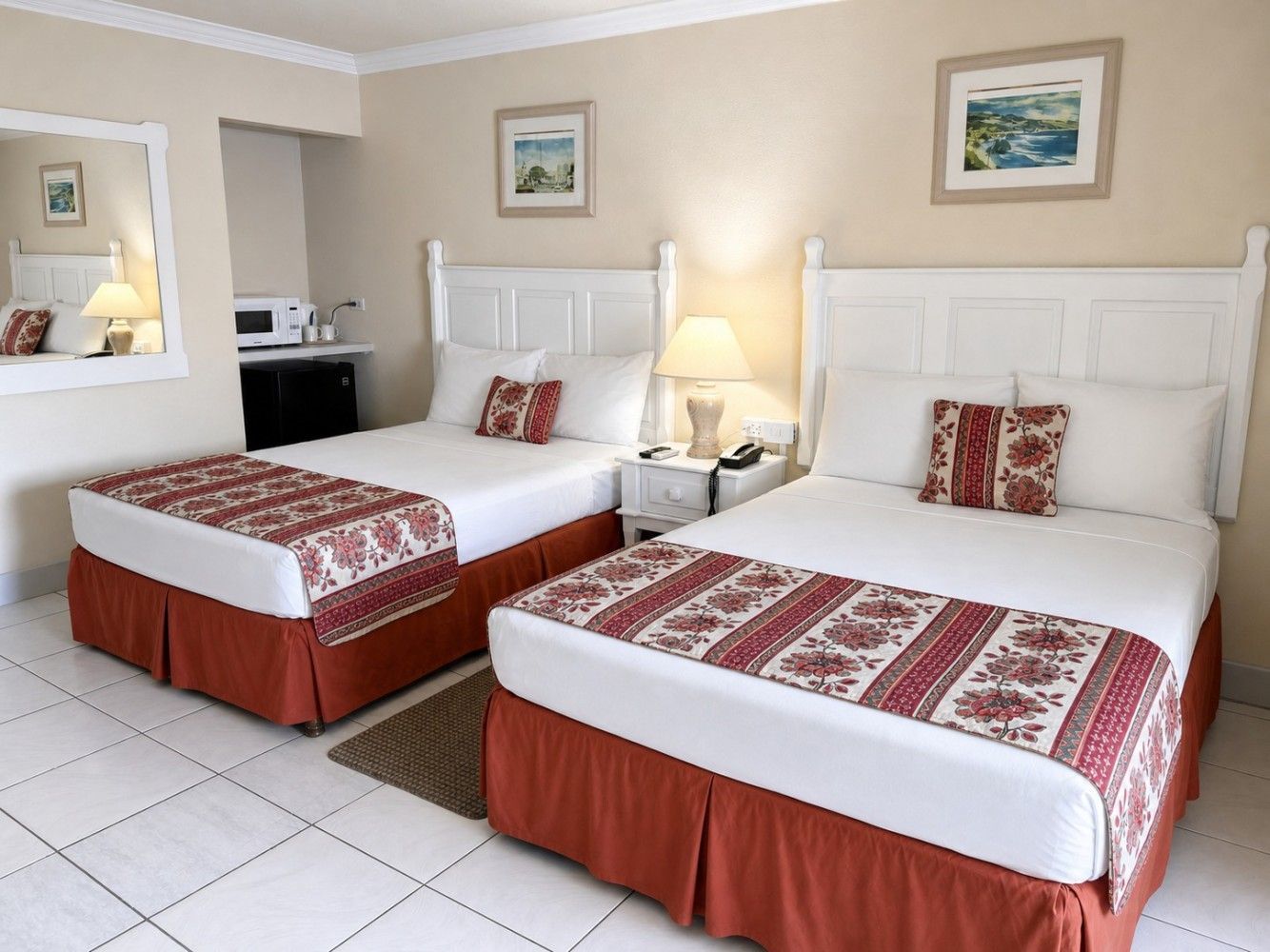 Hotel room with two white beds, red bed skirts, bedside lamp, and framed wall art