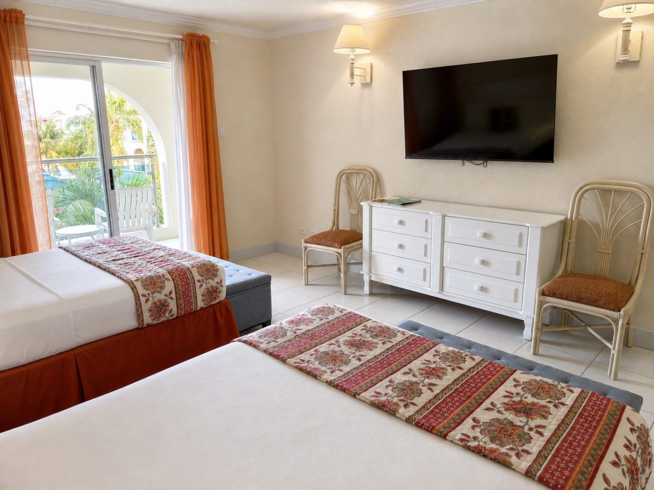 Bright bedroom with two beds, wall-mounted TV, white dresser, wicker chairs, and balcony doors with curtains