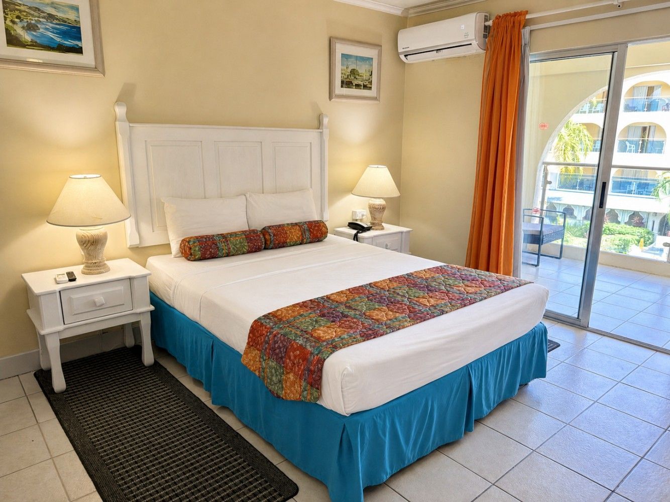 Bright bedroom with white bed, colorful runner, bedside lamps, and balcony doors overlooking a pool.