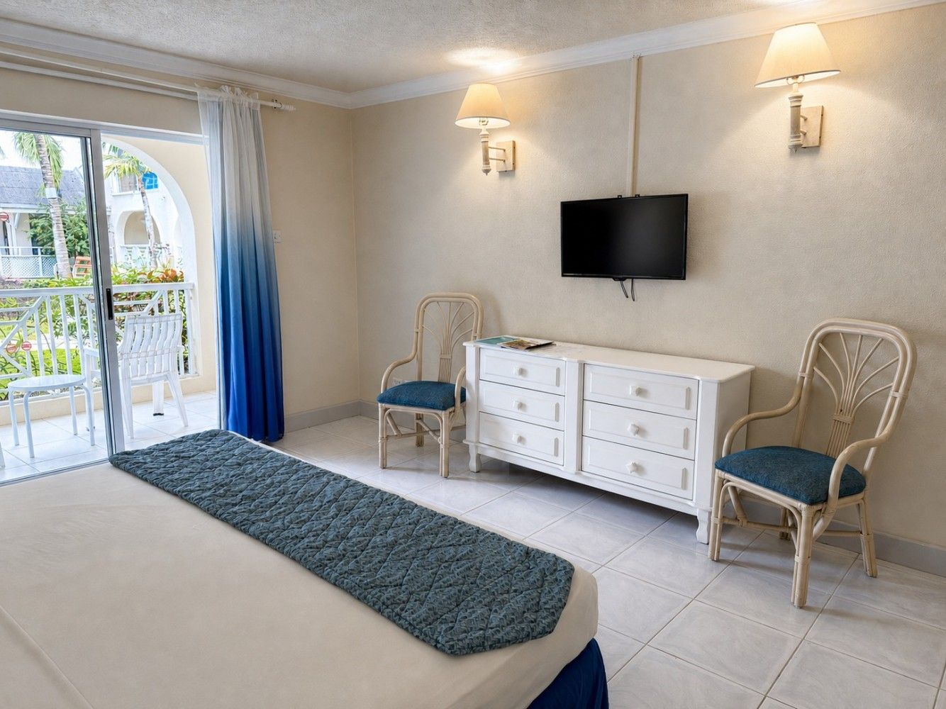 Bright bedroom with twin beds, wall-mounted TV, wicker chairs, and a balcony door with blue curtains.