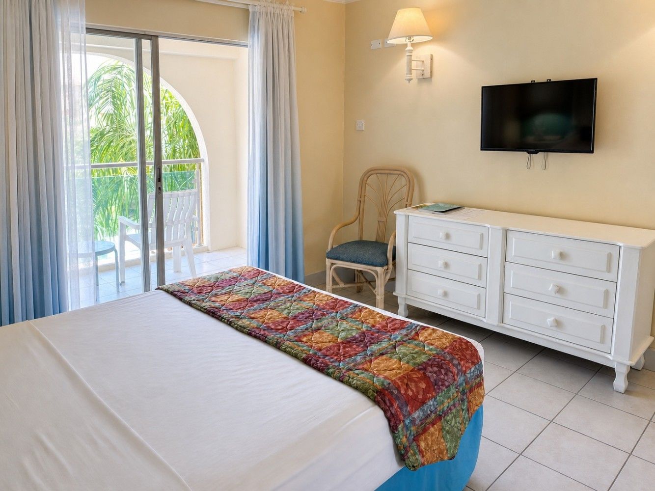 Bright bedroom with a bed, colorful throw, wall-mounted TV, wicker chair, and sliding glass door to a balcony