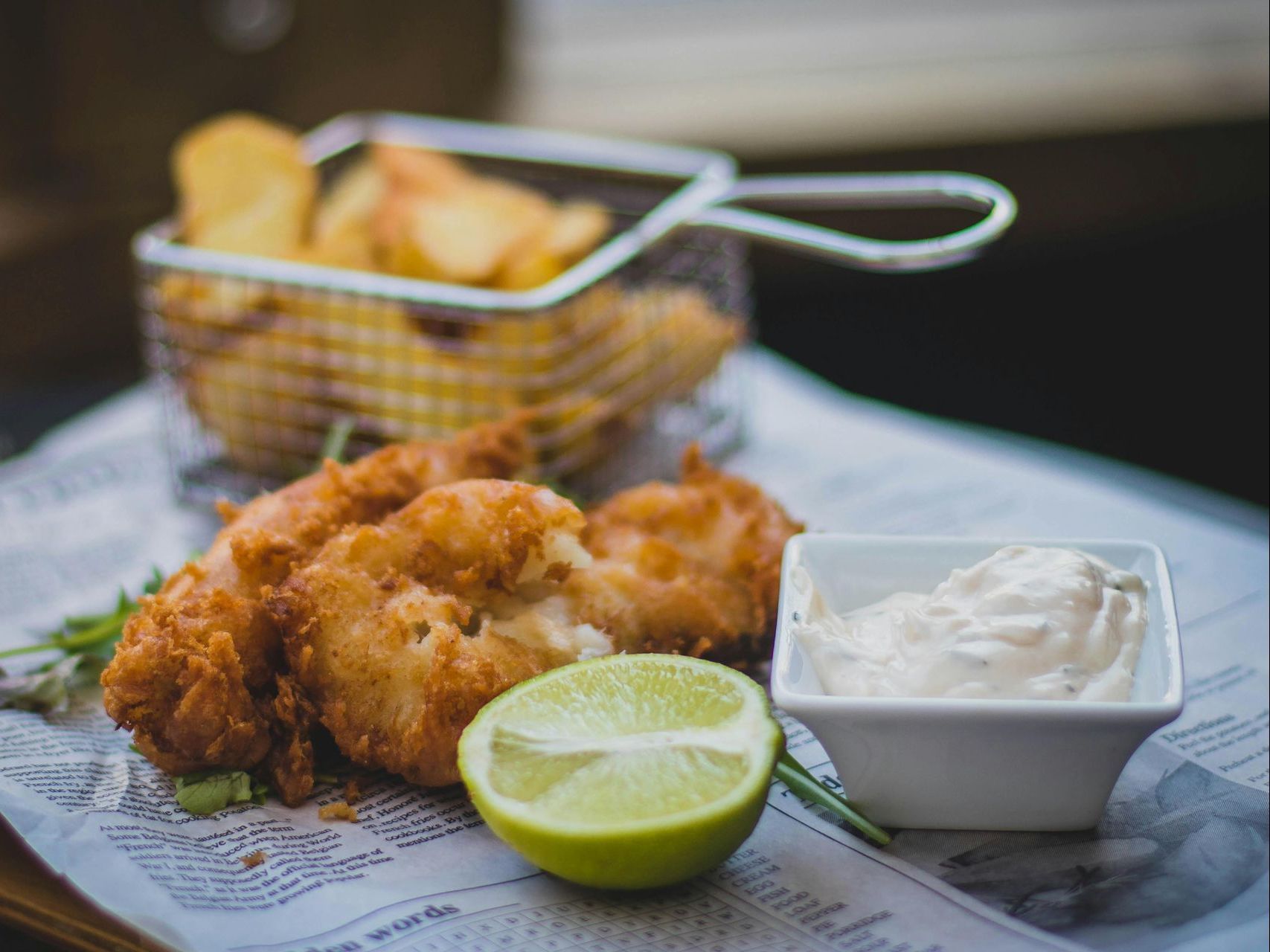 Fried seafood with lime and dipping sauce on newspaper, with fries in a wire basket behind.
