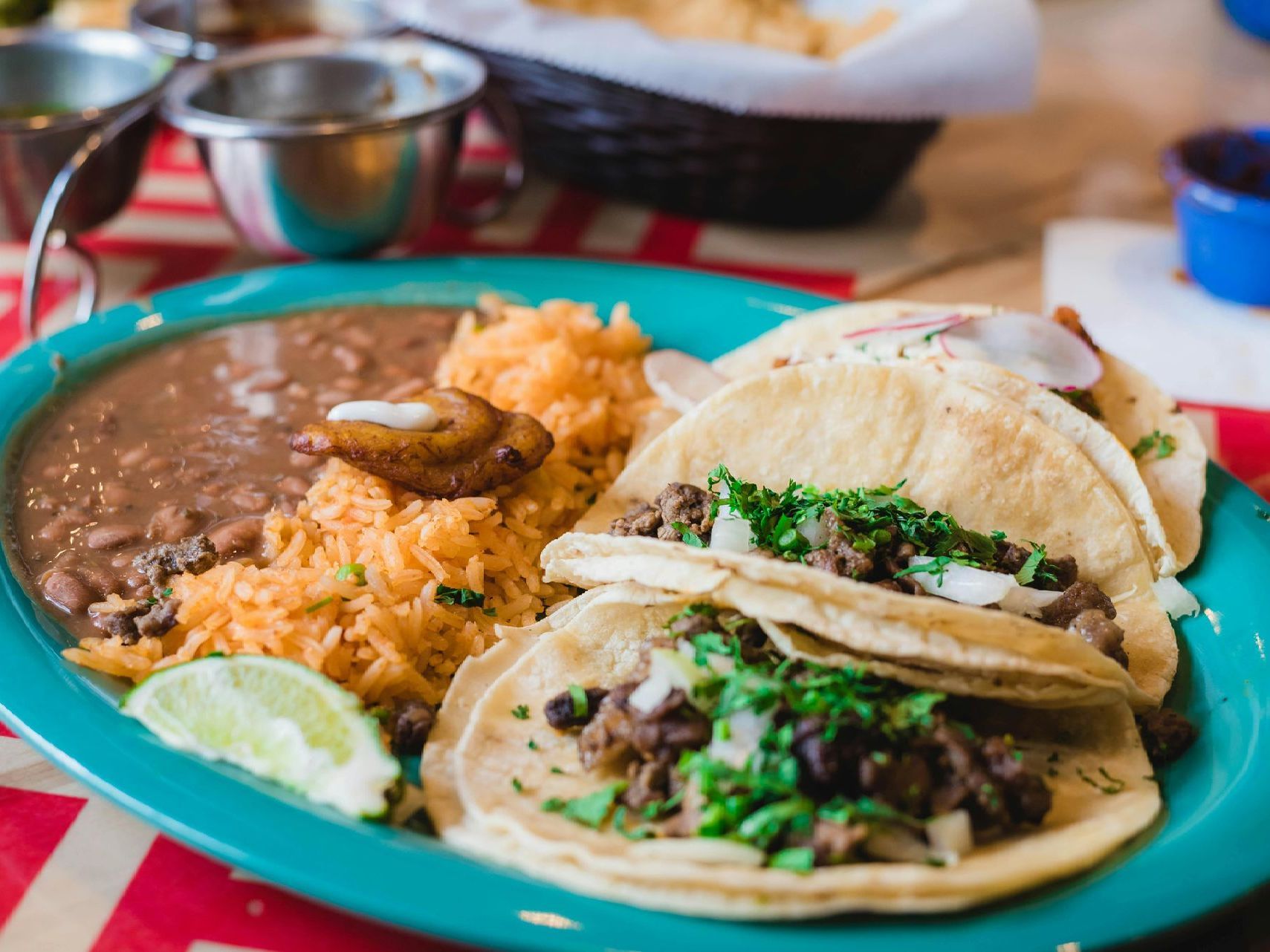 Plate of tacos with rice and beans on a red checkered tablecloth, with salsa and cups in the background