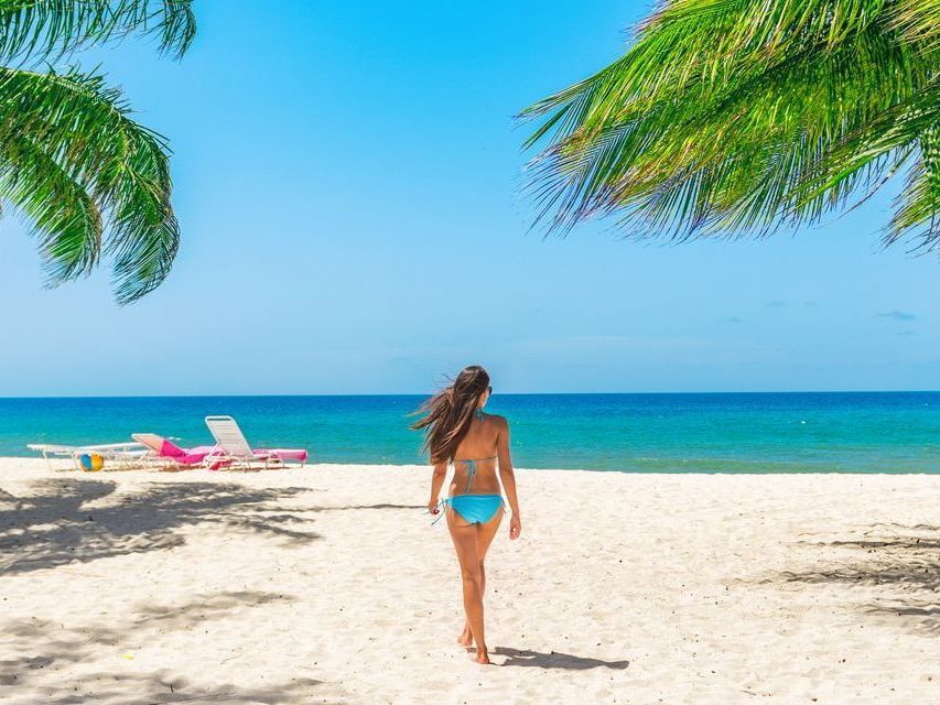 Woman walking on a sunny tropical beach with turquoise water, palm fronds, and lounge chairs.