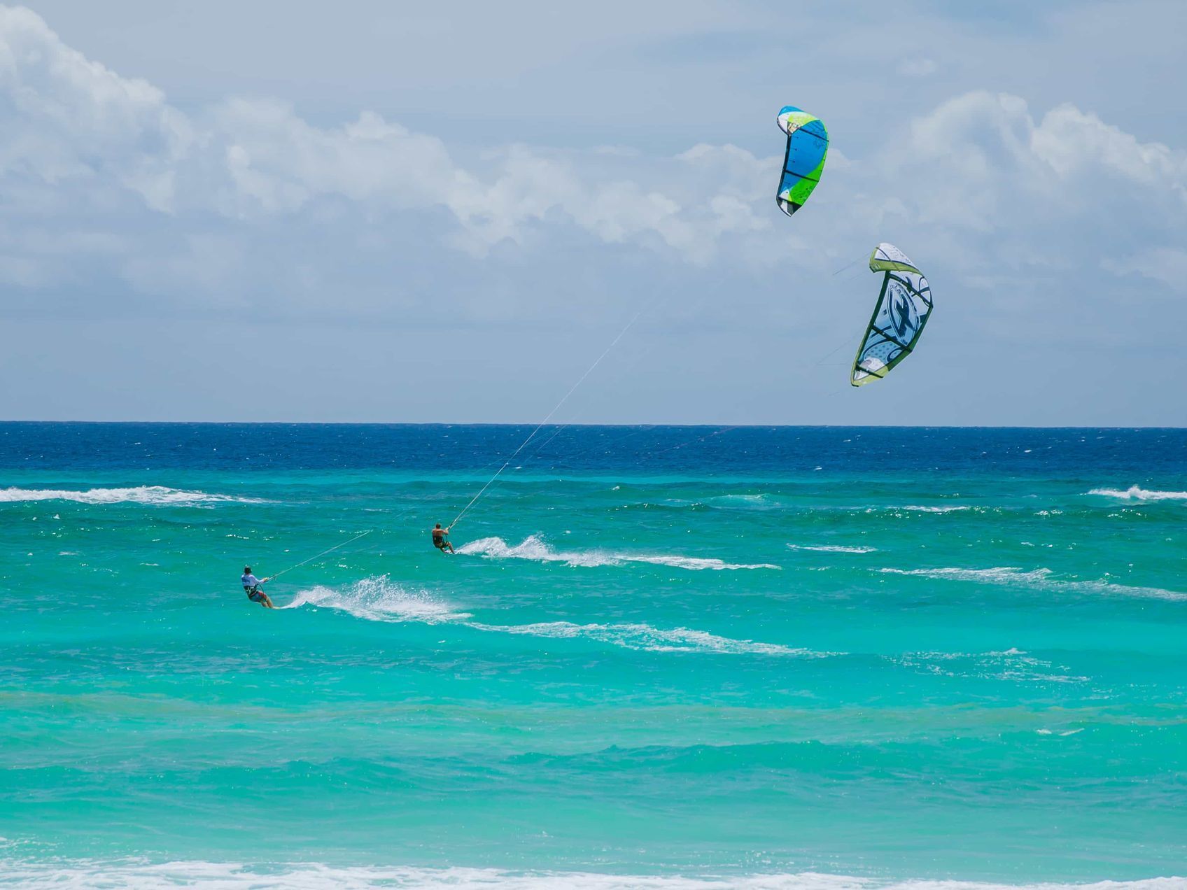 Two kitesurfers glide across turquoise ocean under a cloudy sky