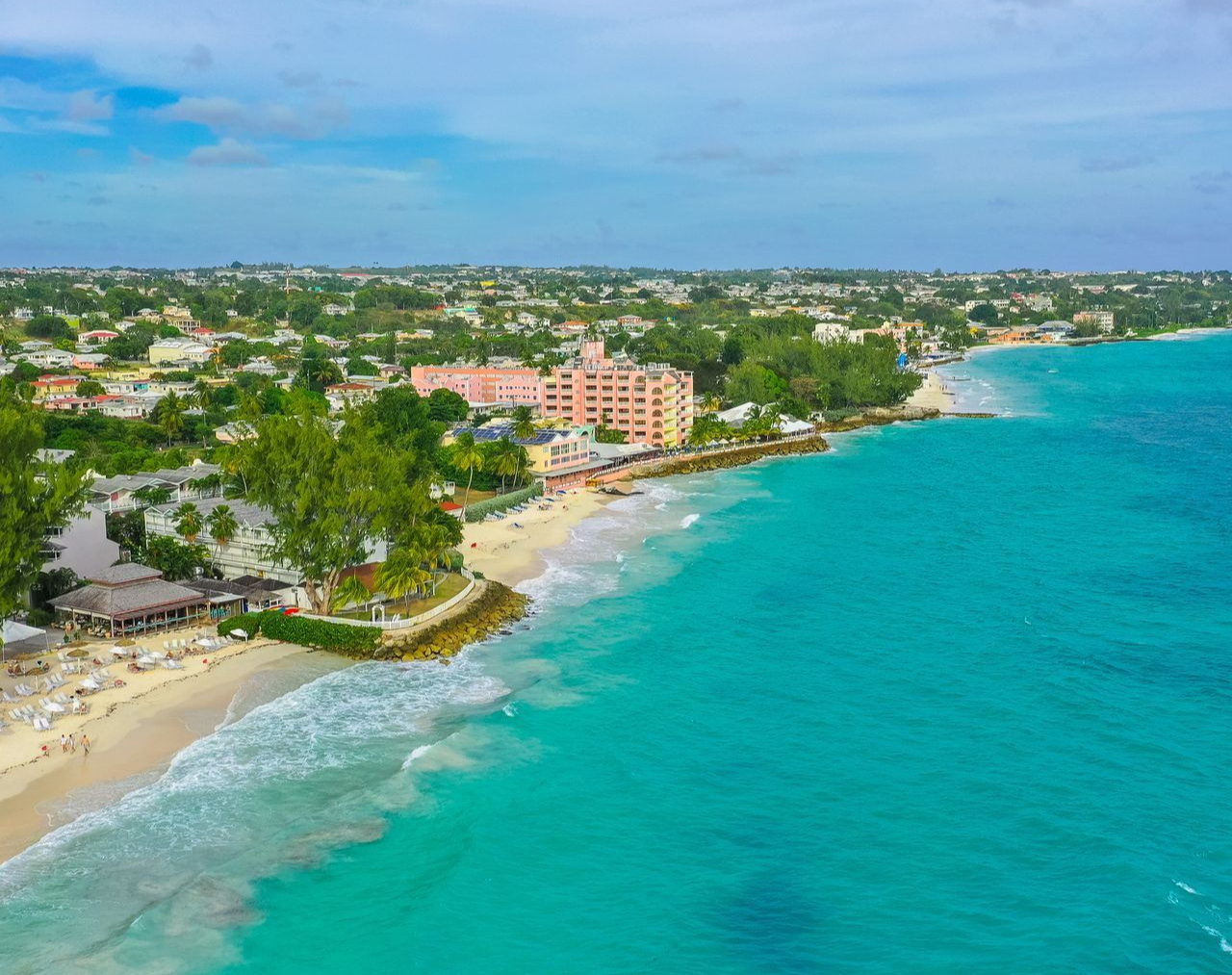 Aerial view of a turquoise beach with white surf, palm trees, and seaside buildings along the shore