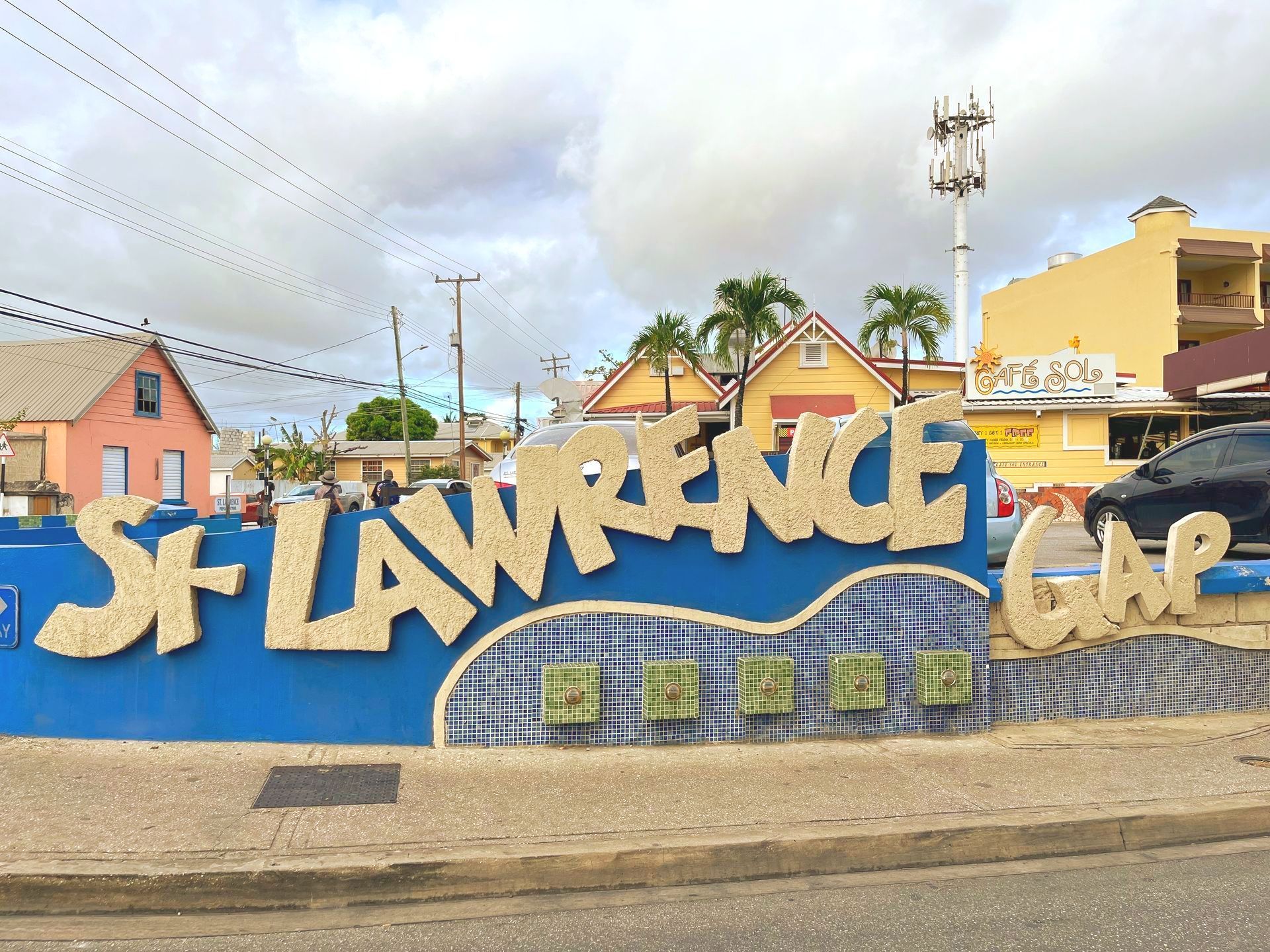 Blue graffiti-style “ST LAURENCE” mural on a stone wall in a suburban street with houses and a cloudy sky