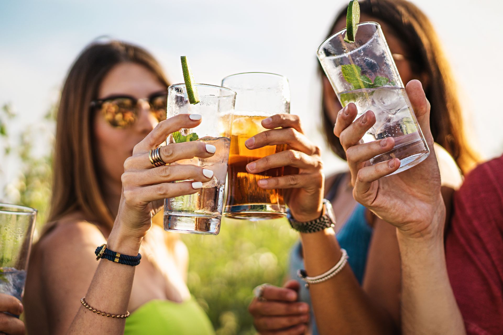 Friends clink cocktails and iced drinks outdoors in a sunny garden setting.
