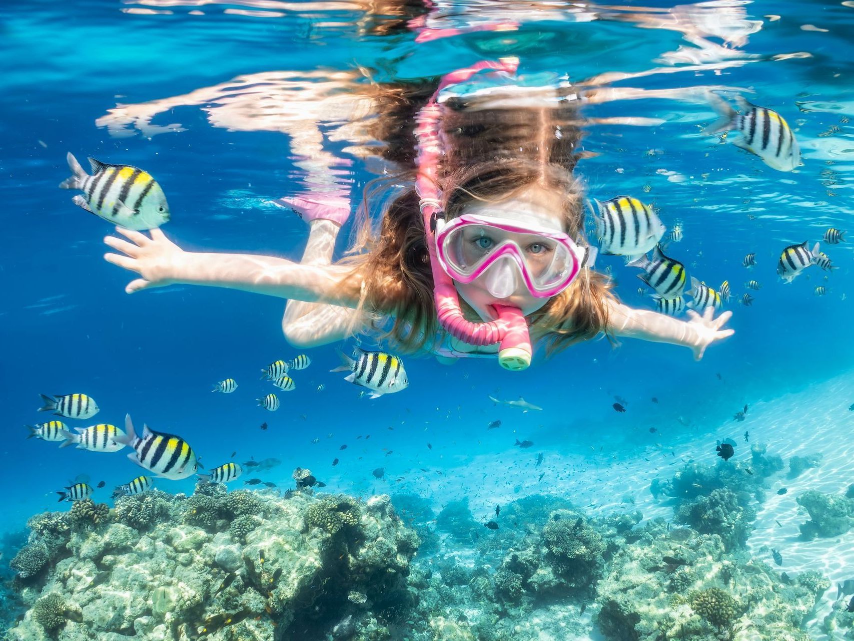 Child snorkeling over coral reef with pink mask, surrounded by striped fish in clear blue water