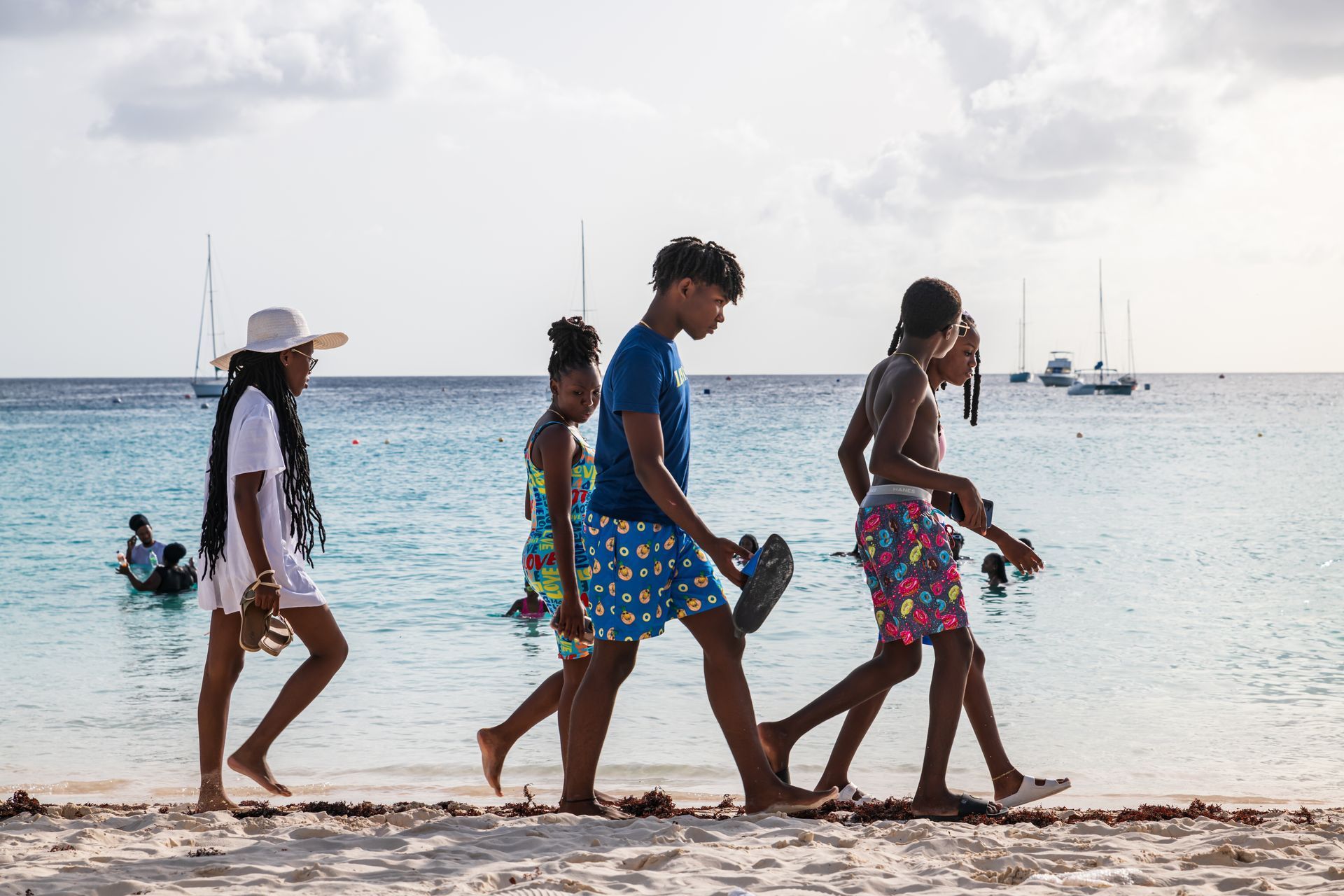 Four people walking along a sandy beach beside calm water, with sailboats in the distance.