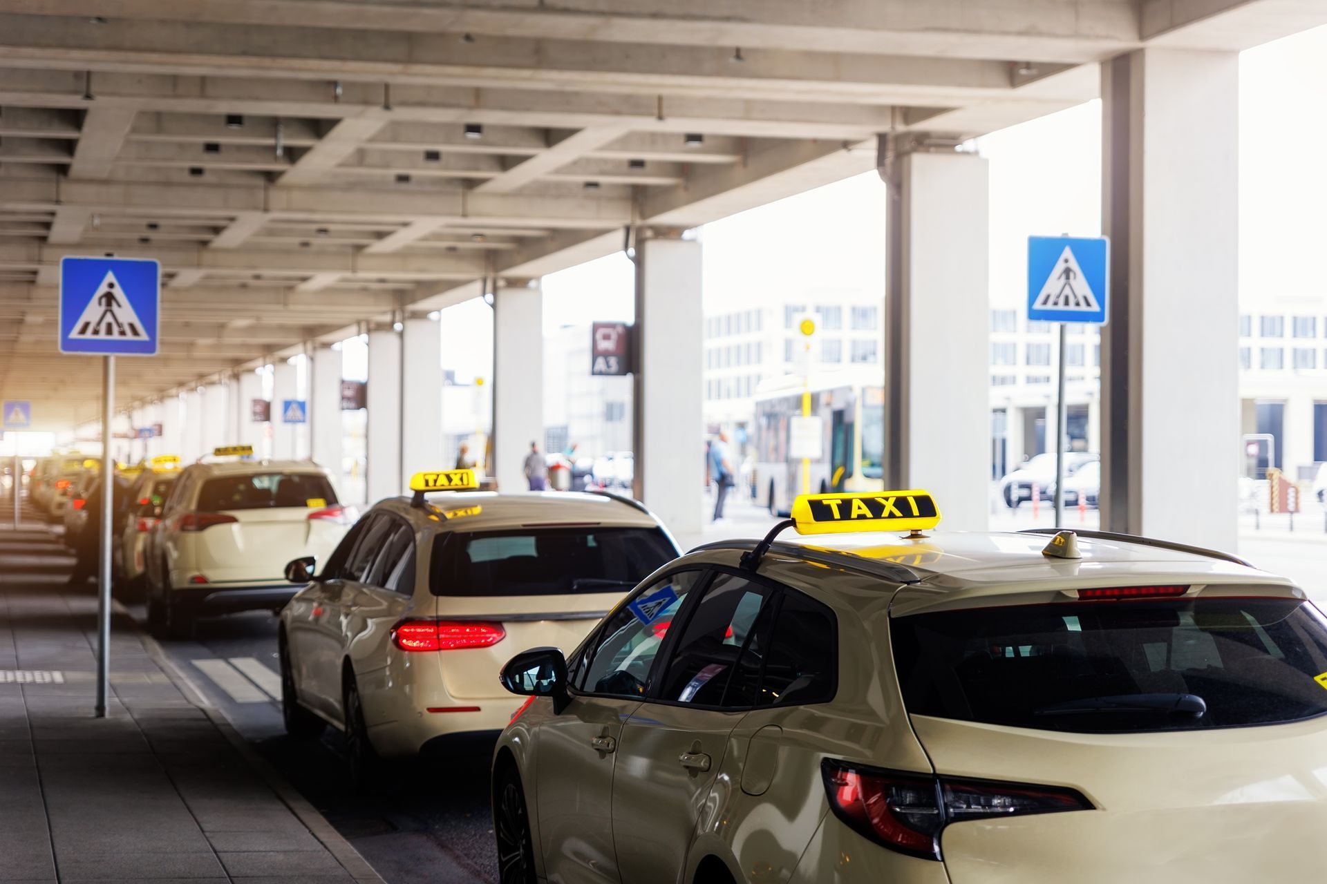 Taxis waiting in a covered airport pickup lane with blue pedestrian crossing signs.