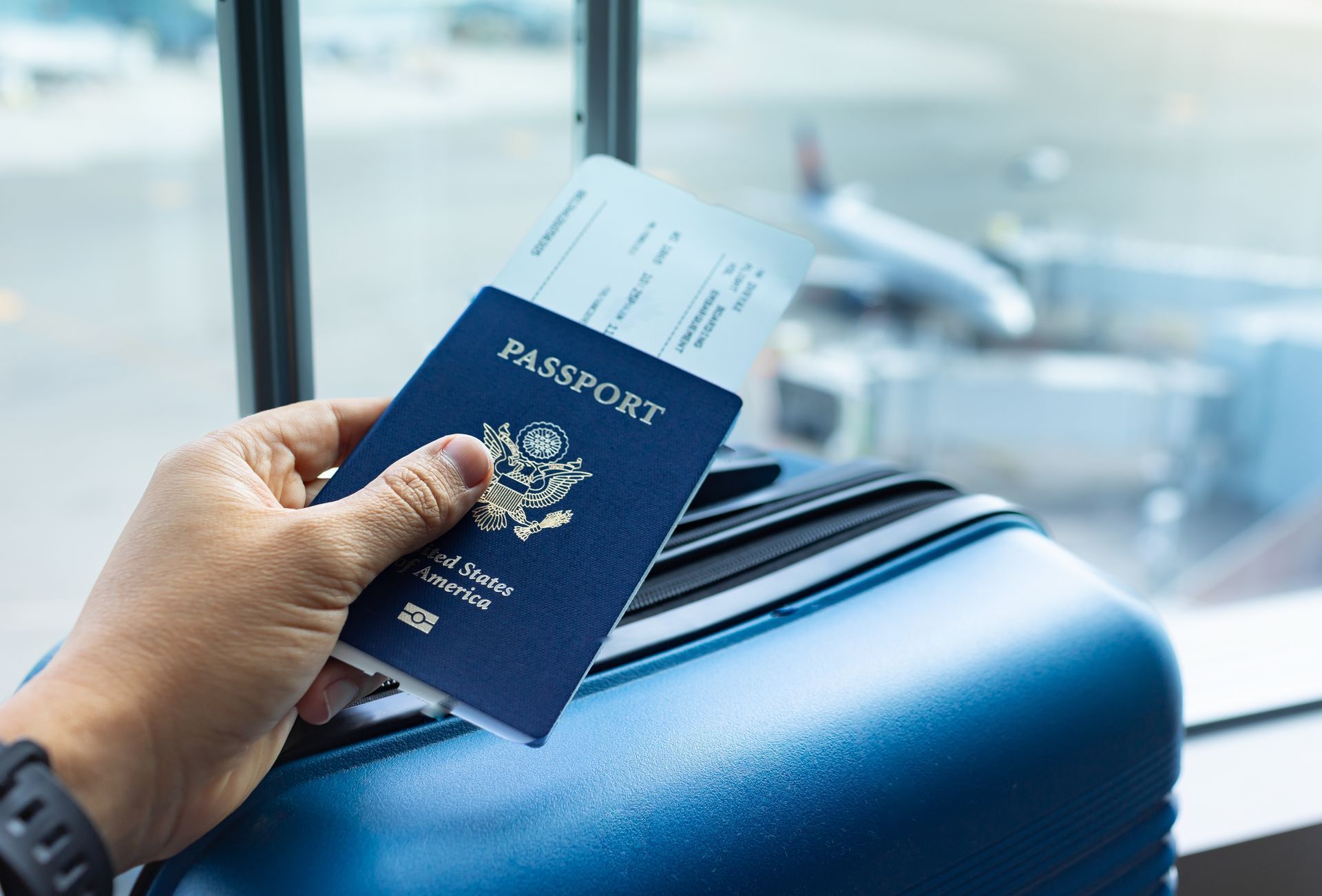 Hand holding a blue passport beside a suitcase at an airport window